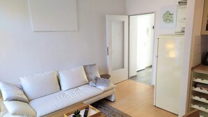 Ein modernes Wohnzimmer mit einem weißen Sofa, einem Holztisch und einer kleinen Pflanze. Eine Tür führt zu einem Flur mit einem Kühlschrank und einem Bild von Paris.