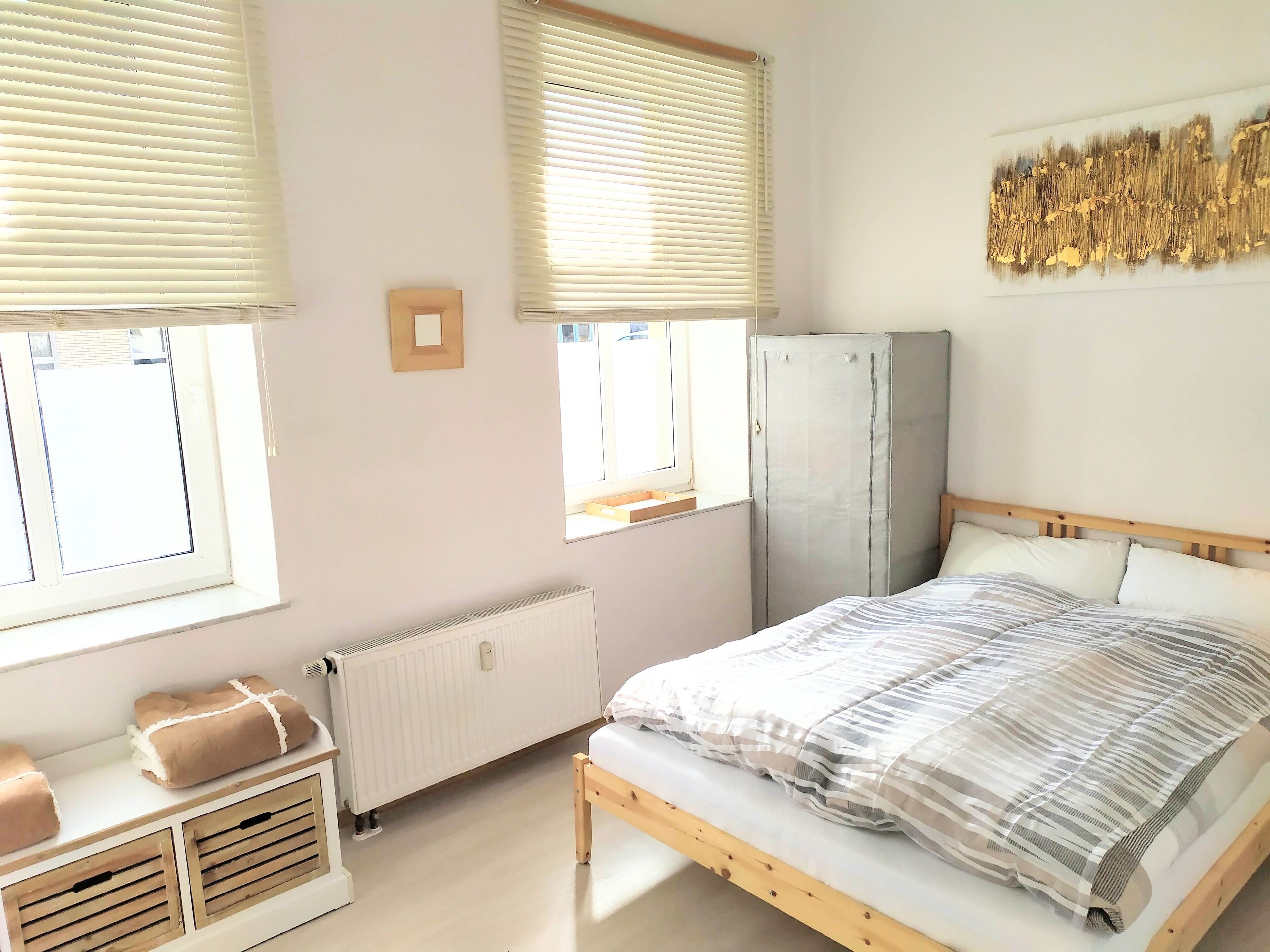 A bedroom with a bed, a radiator, a wardrobe, and two windows with blinds.