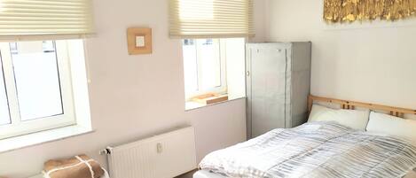 A bedroom with a bed, a radiator, a wardrobe, and two windows with blinds.