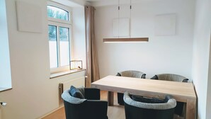 Un espace repas moderne avec une table en bois et des chaises noires dotées de coussins gris. On y trouve une fenêtre avec des rideaux, un luminaire suspendu et un radiateur au mur.