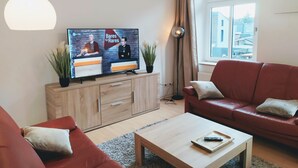 Un salon moderne avec un téléviseur, des canapés rouges, une table basse en bois et une vue sur la ville par une fenêtre.