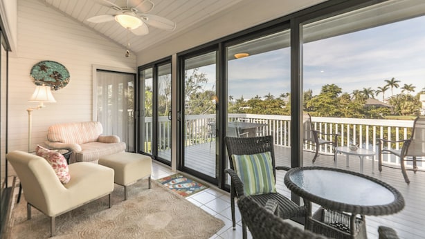 Un balcon spacieux doté d’un ventilateur de plafond, un coin salon confortable avec une table, et une vue sur des palmiers et une côte lointaine.