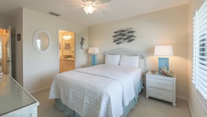 Une chambre à coucher comprenant un lit, deux tables de chevet avec des lampes, un ventilateur de plafond, un miroir fixé au mur et une décoration murale représentant des poissons.