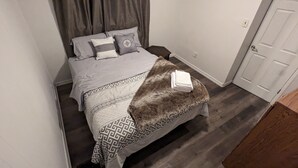 Een slaapkamer met een bed, grijs beddengoed, gestreepte kussens, een zachte plaid en een klein nachtkastje met een lamp.