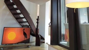 Tilava huone, jossa on puinen portaikko, suuri ikkuna, josta on kaunis näköala, ja televisio, jolla näkyy auringonlaskumaisema.