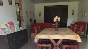 Een eetruimte met een grote tafel, bedekt met een tafelkleed met bloemenmotief, omringd door houten stoelen. Links bevindt zich een kast met decoratieve objecten.
