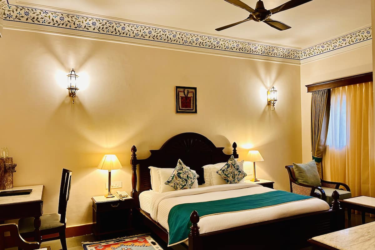 A hotel room with a bed, two bedside tables with lamps, a chair, a desk, a ceiling fan, and a window with curtains.