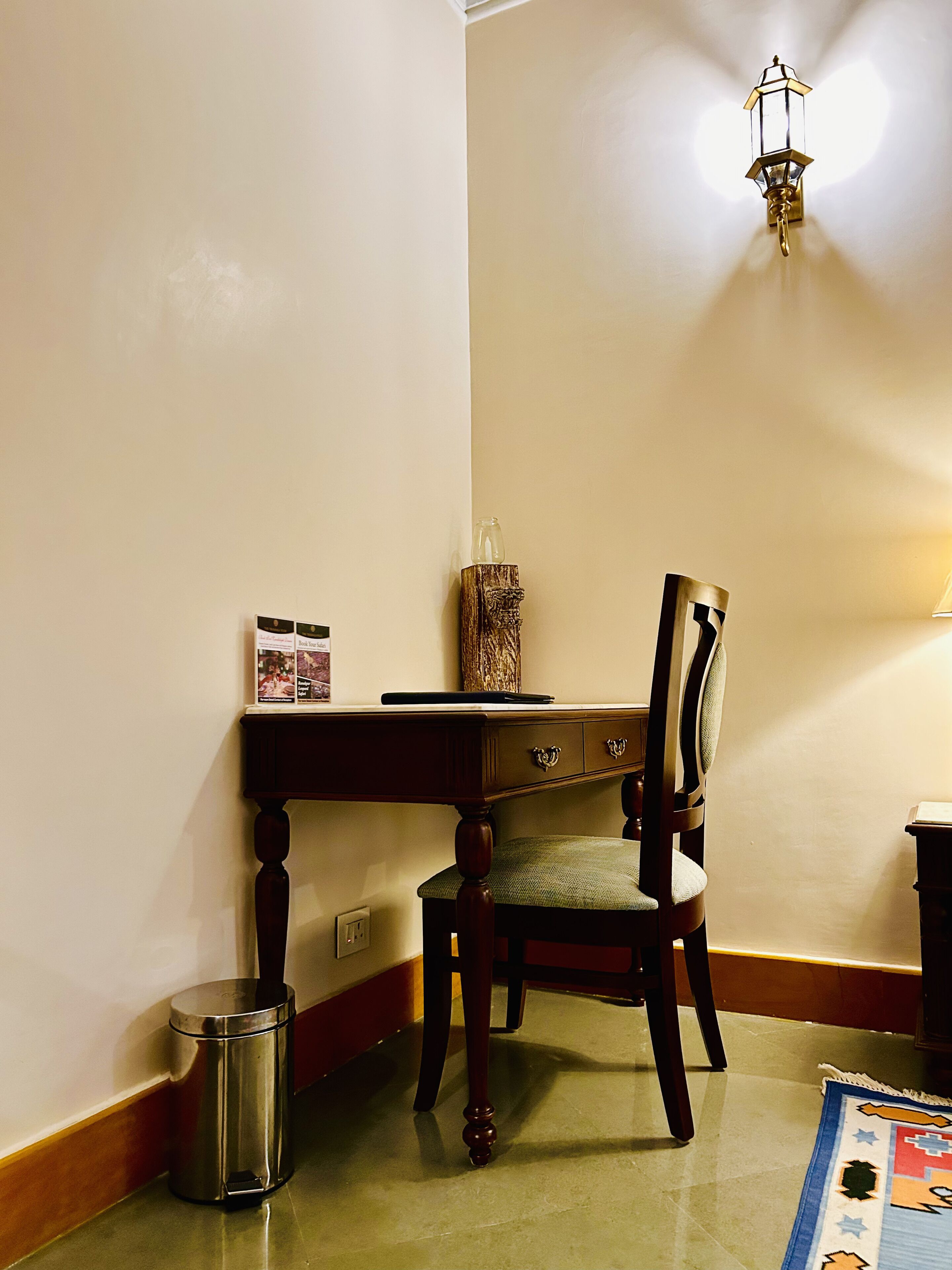 A corner room with a wooden table, a chair, a trash bin, and a wall-mounted light fixture.