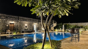 A nighttime swimming pool with a stone wall backdrop, lounge chairs, and a small tree. - The Trishala Vilas Hotel in Ranakpur (Desuri)