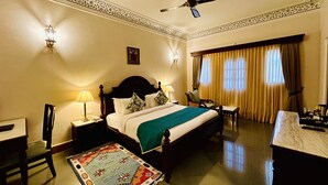 A hotel room with a large bed, a desk, a chair, a ceiling fan, and a window with curtains. - The Trishala Vilas Hotel in Ranakpur (Desuri)