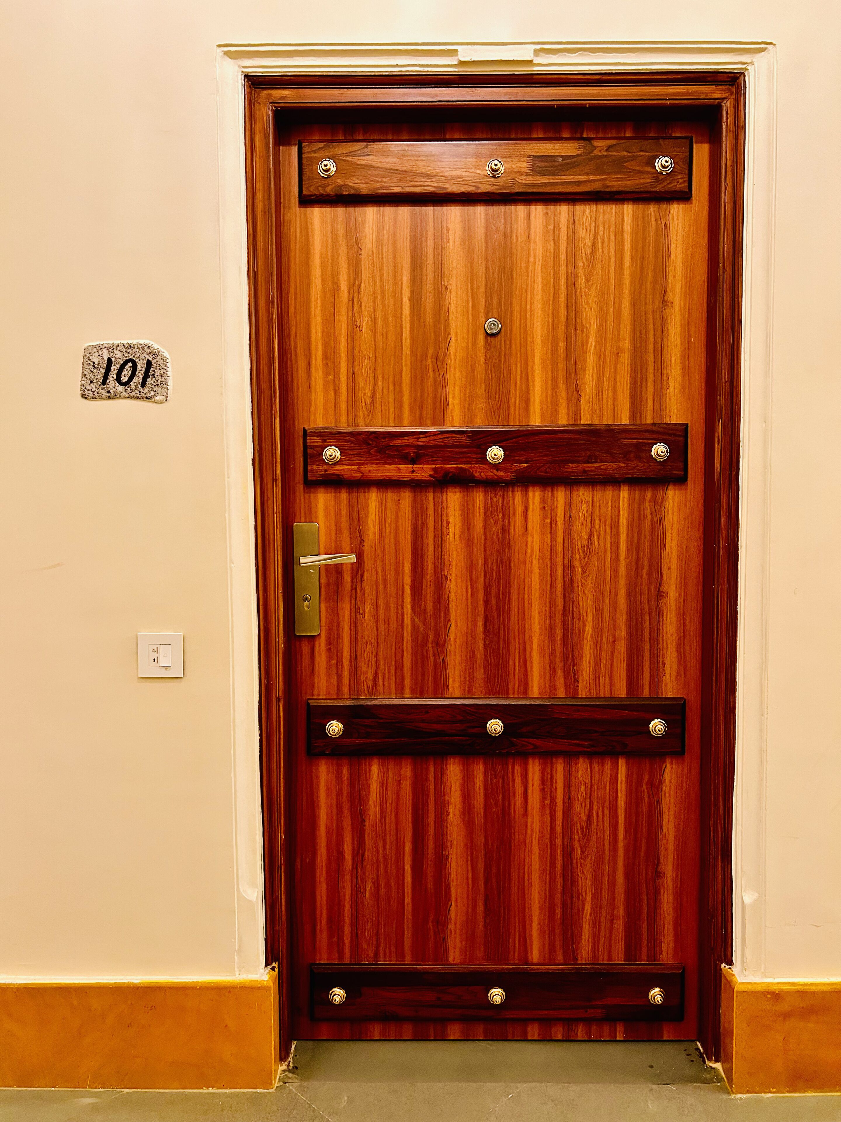 Una puerta de madera con picaporte de latón, una placa con número y un interruptor de luz en la pared.