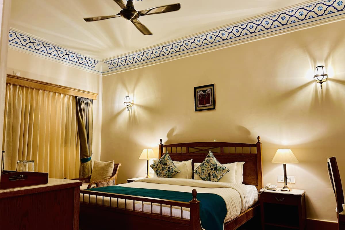 A hotel room with a wooden bed, two bedside lamps, a ceiling fan, a wall-mounted picture, and a window with curtains.