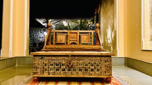 A carved wooden chest with intricate designs, placed on a patterned rug in a hallway with a brick wall and a palm tree in the background. - The Trishala Vilas Hotel in Ranakpur (Desuri)