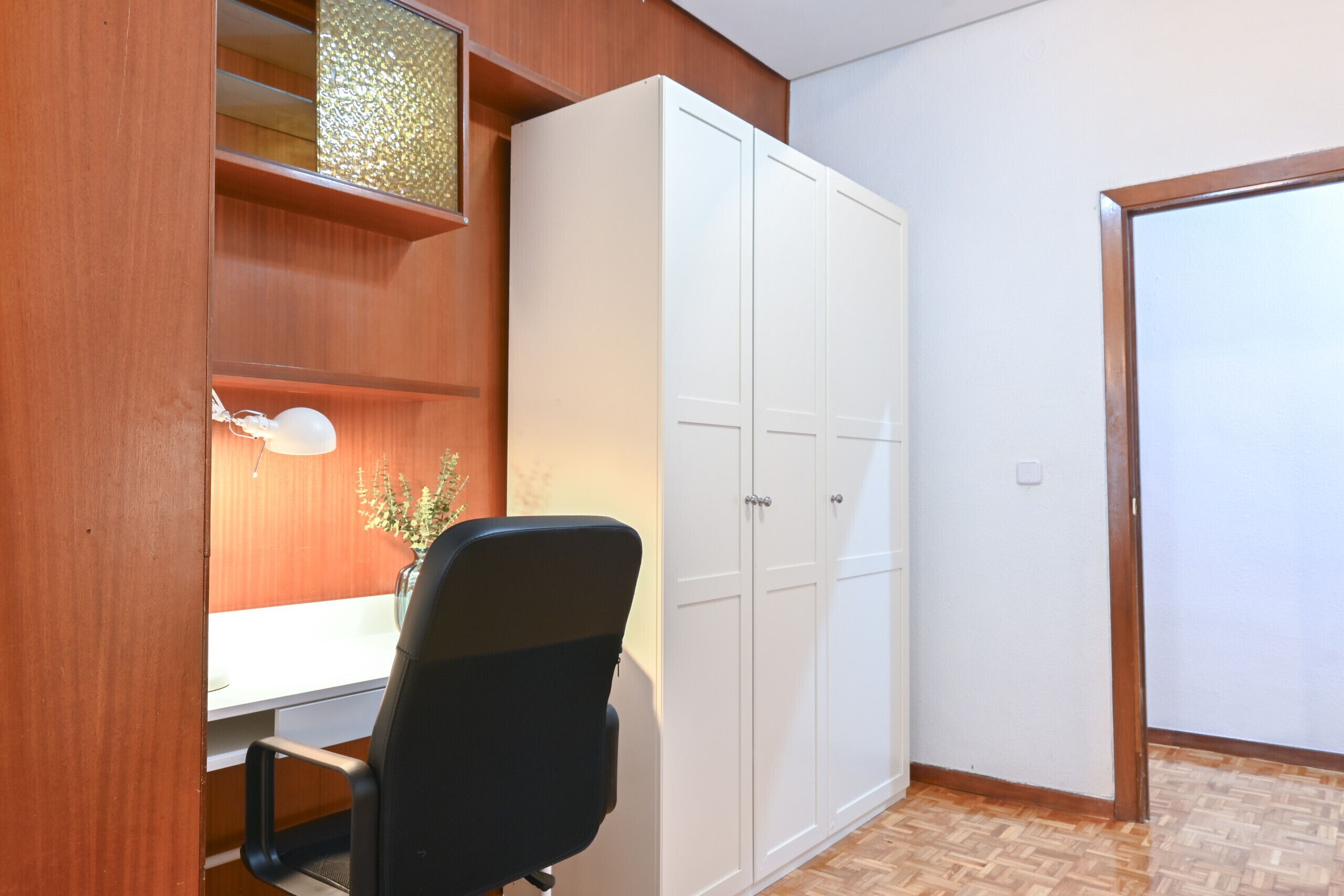 A room with a wooden desk, a black office chair, a white wardrobe, and a wooden floor.