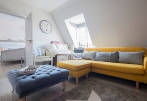 A modern living room with a yellow sofa, a blue ottoman, and a white chair. A clock is on the wall, and there is a window with a view of the outside. - The Loft | Exposed Brick & Fire | Cowdray Views | South Downs | Midhurst High St (Midhurst)