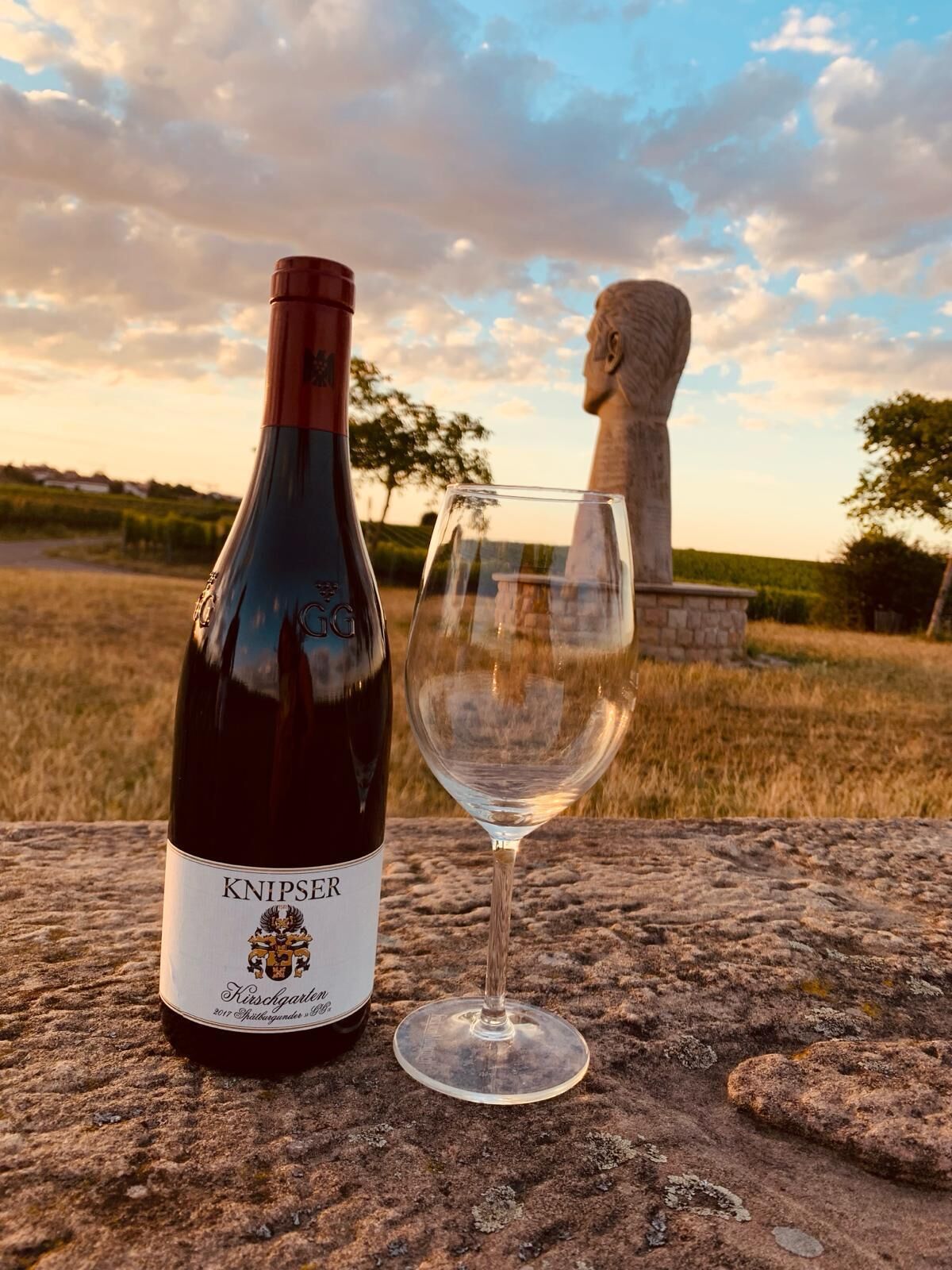Una botella de vino Knipper y una copa vacía sobre una superficie de piedra con una escultura al fondo.