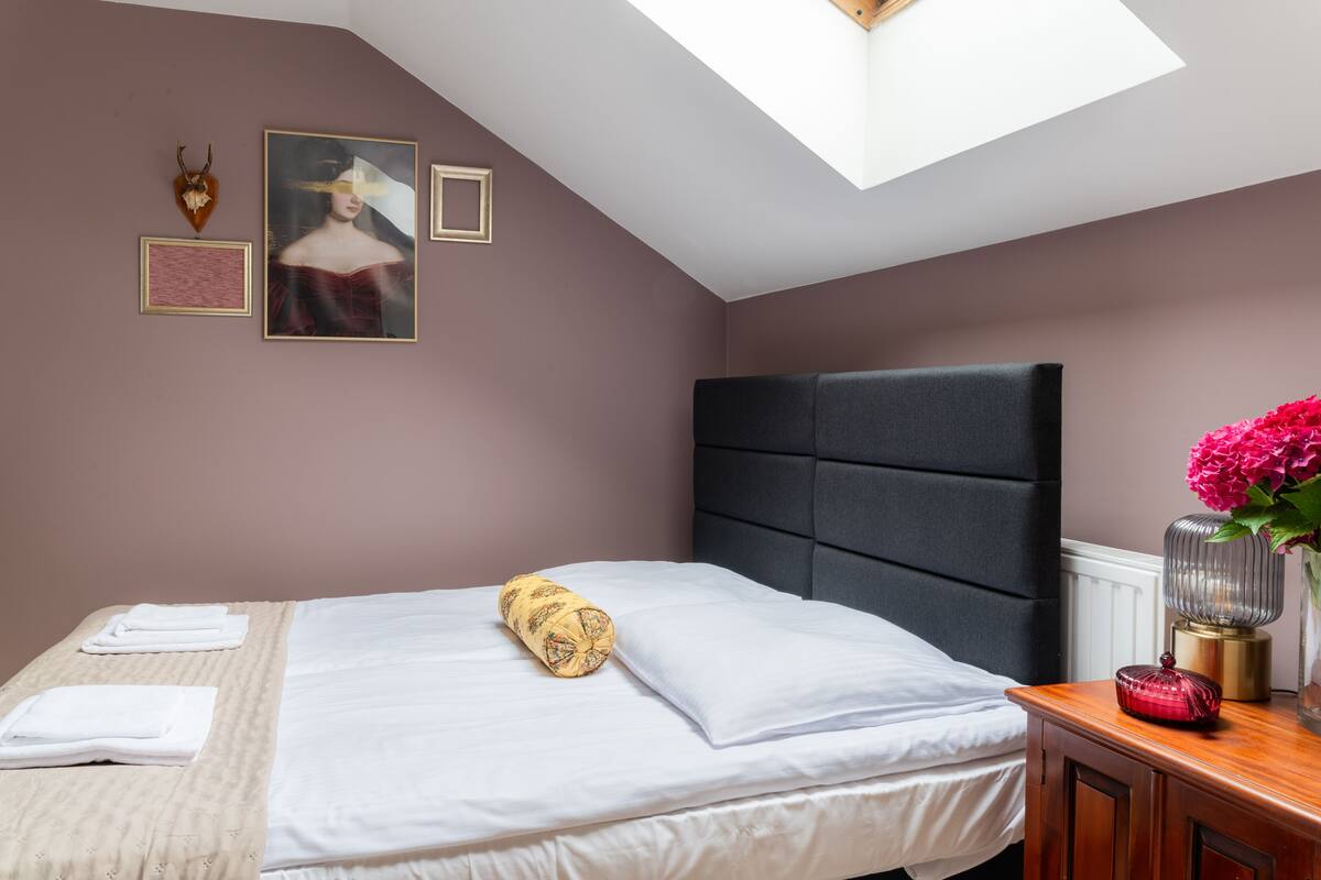 A neatly made bed with white linens and a rolled towel, a dark upholstered headboard, and a wooden nightstand with a vase of flowers.