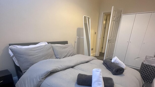 A neatly made bed with grey linens and rolled towels, a bedside table, a wardrobe, and a mirror.