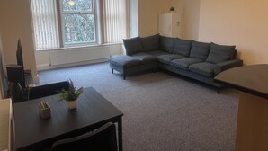 A living room with a grey sectional sofa, a small table with a plant, and a window with blinds.
