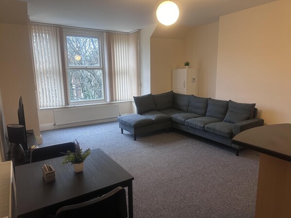 A living room with a grey sectional sofa, a small table with a plant, and a window with blinds.