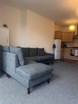 A living room with a grey sectional sofa, a white refrigerator, wooden cabinets, and a tiled kitchen area.