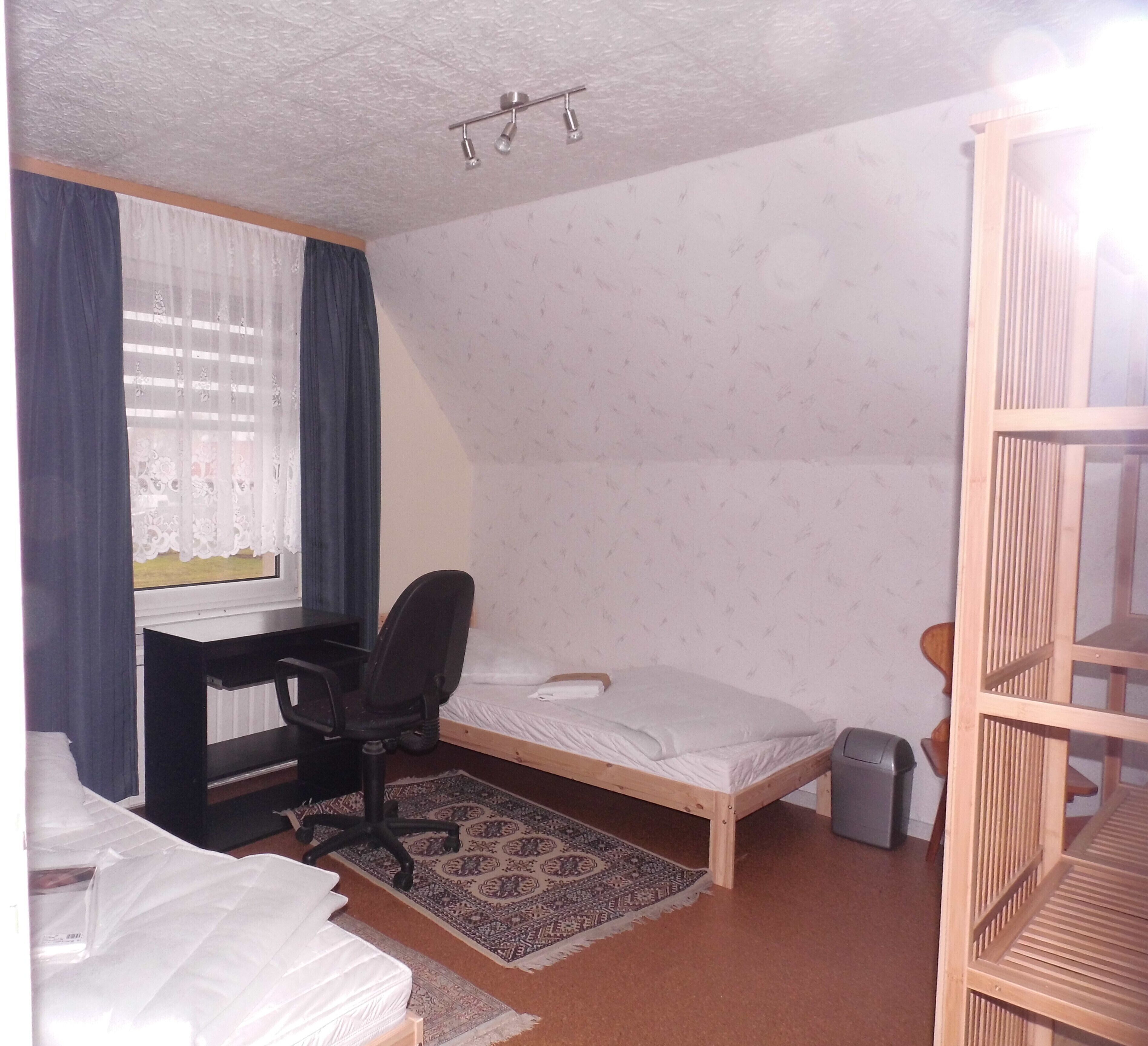 A small, compact room with a single bed, a desk with a chair, a small rug, and a window with curtains.
