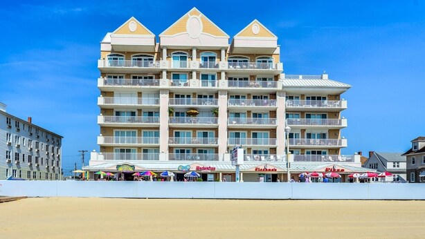 Un hôtel à plusieurs étages doté de balcons, situé en bord de mer et comprenant plusieurs boutiques, dont une pizzeria.