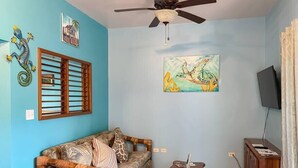 Un salon avec un mur bleu, un canapé en bois, un ventilateur de plafond, un tableau représentant une tortue et un téléviseur posé sur un meuble en bois.