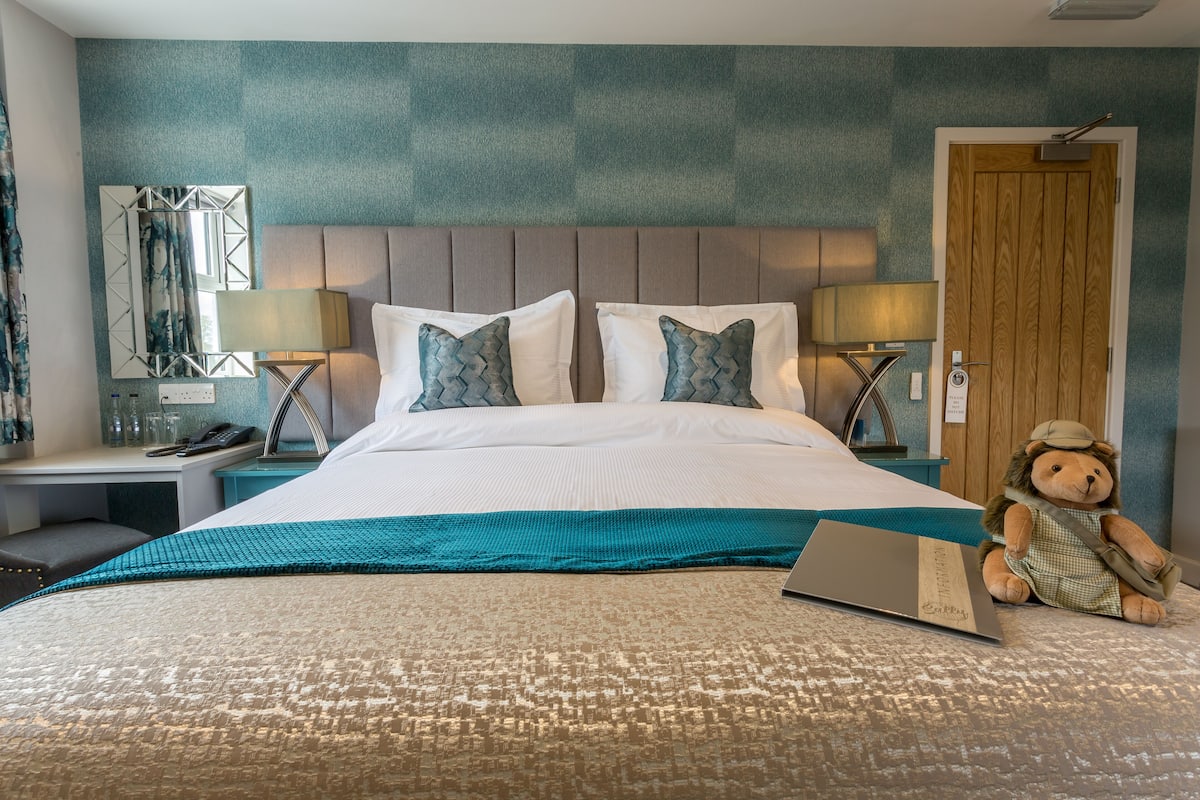 A neatly made bed with a teal throw, a laptop, and a teddy bear on the bed.