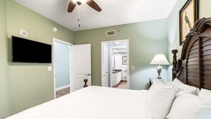 Un dormitorio con una cama grande, un ventilador de techo, un televisor de pantalla plana montado en la pared y una puerta que da a un pasillo.