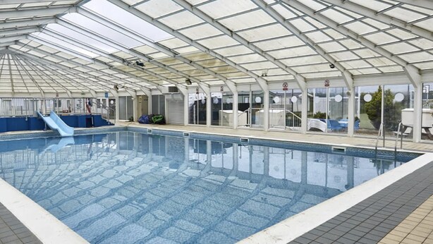 Une piscine intérieure dotée d’un toit en verre, d’un toboggan et d’un espace aménagé au bord du bassin avec des sièges.
