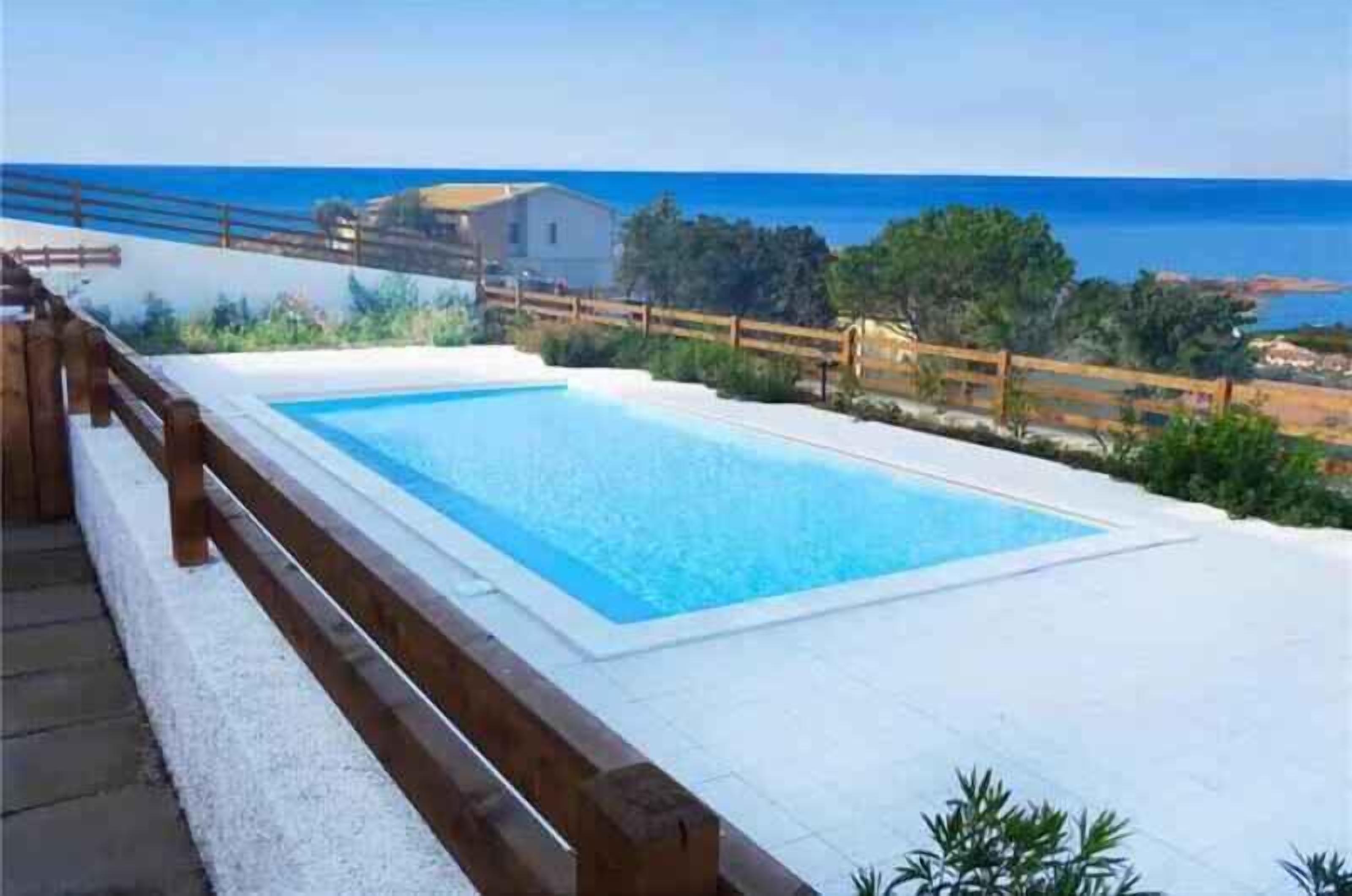 En rektangulär swimmingpool med ett trädäck och ett räcke runt om, omgiven av grönska och med utsikt över havet.
