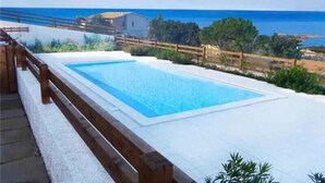 Ein rechteckiger Pool mit Holzterrasse und Geländer, umgeben von Grünpflanzen und Blick aufs Meer.
