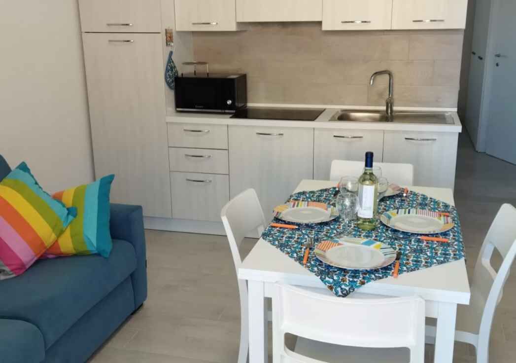 Una cocina moderna con armarios blancos, un microondas negro y un lavamanos. Una mesa de comedor con mantel colorido está servida con platos y cubiertos.