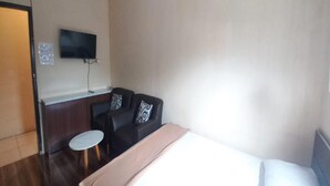 A hotel room with a bed, a small round table, two black sofas with patterned cushions, a television mounted on the wall, and a door. - OYO 93668 Buah Batu Guest House (Bandung)