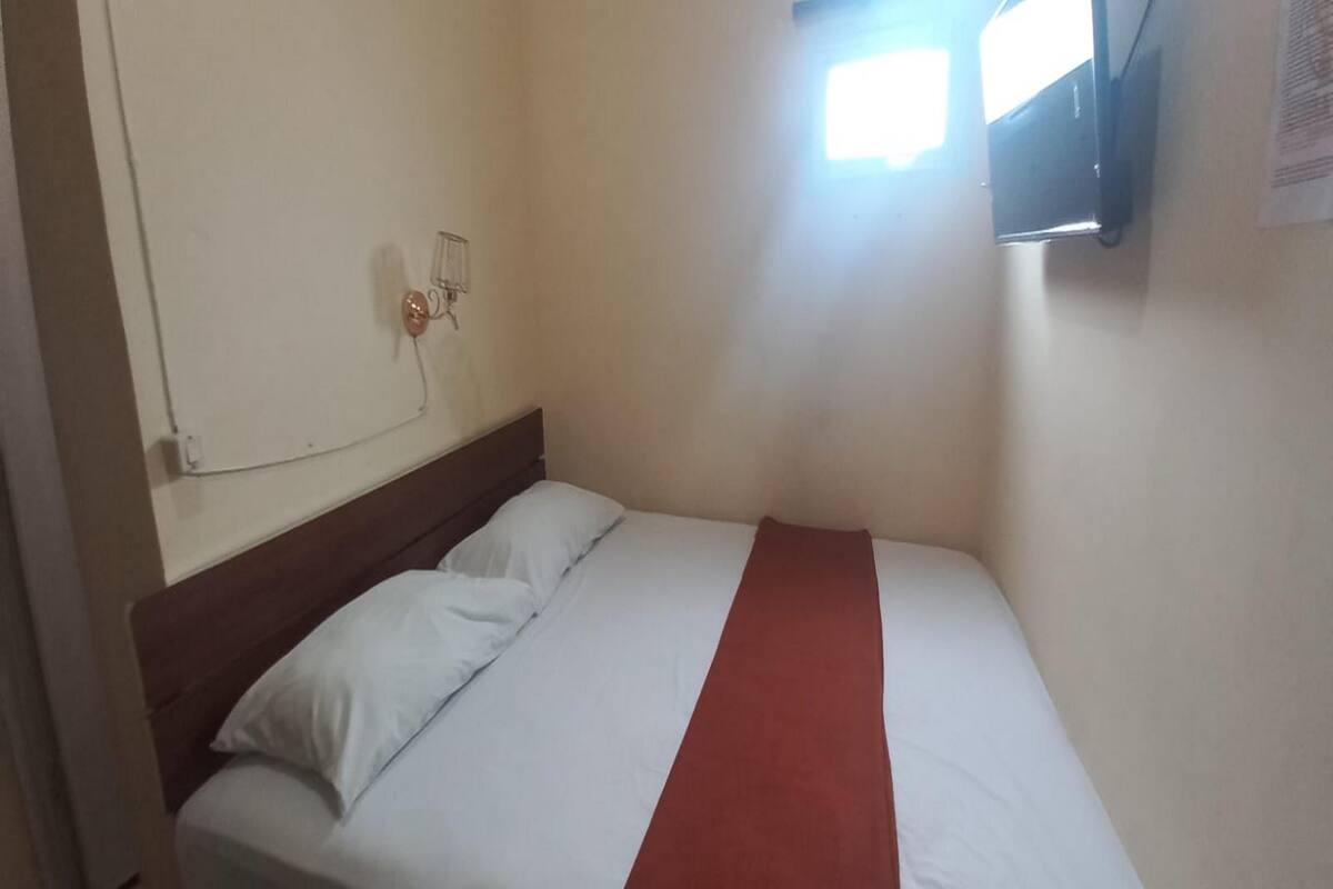 A small, single-bed room with a wooden headboard, white bedding, and a red accent.
