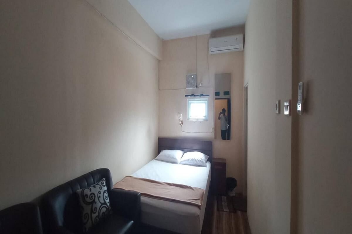 A small, single-bed room with a bed, a bedside table, a chair, a window, and an air conditioning unit.