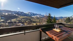 Un balcón con una mesa de madera y dos copas de vino tinto, con vistas a un paisaje montañoso de picos nevados y cielo despejado.