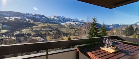 A balcony with a wooden table and two glasses of red wine, overlooking a mountainous landscape with snow-capped peaks and a clear sky.