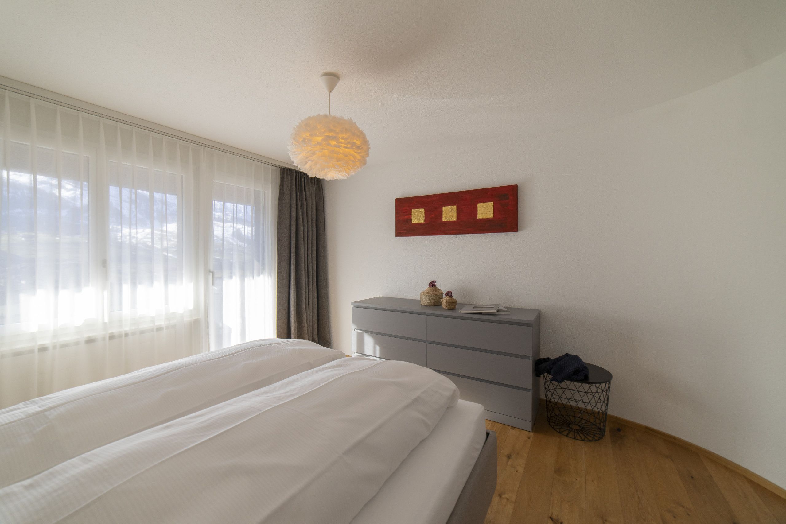 Un dormitorio con cama, cómoda, lámpara de pared y vista a través de una ventana con cortinas translúcidas.