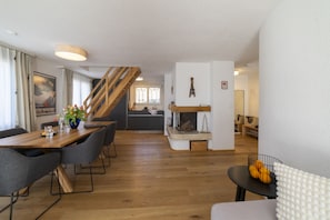 A modern interior with a wooden dining table, grey chairs, a staircase, and a kitchen area.