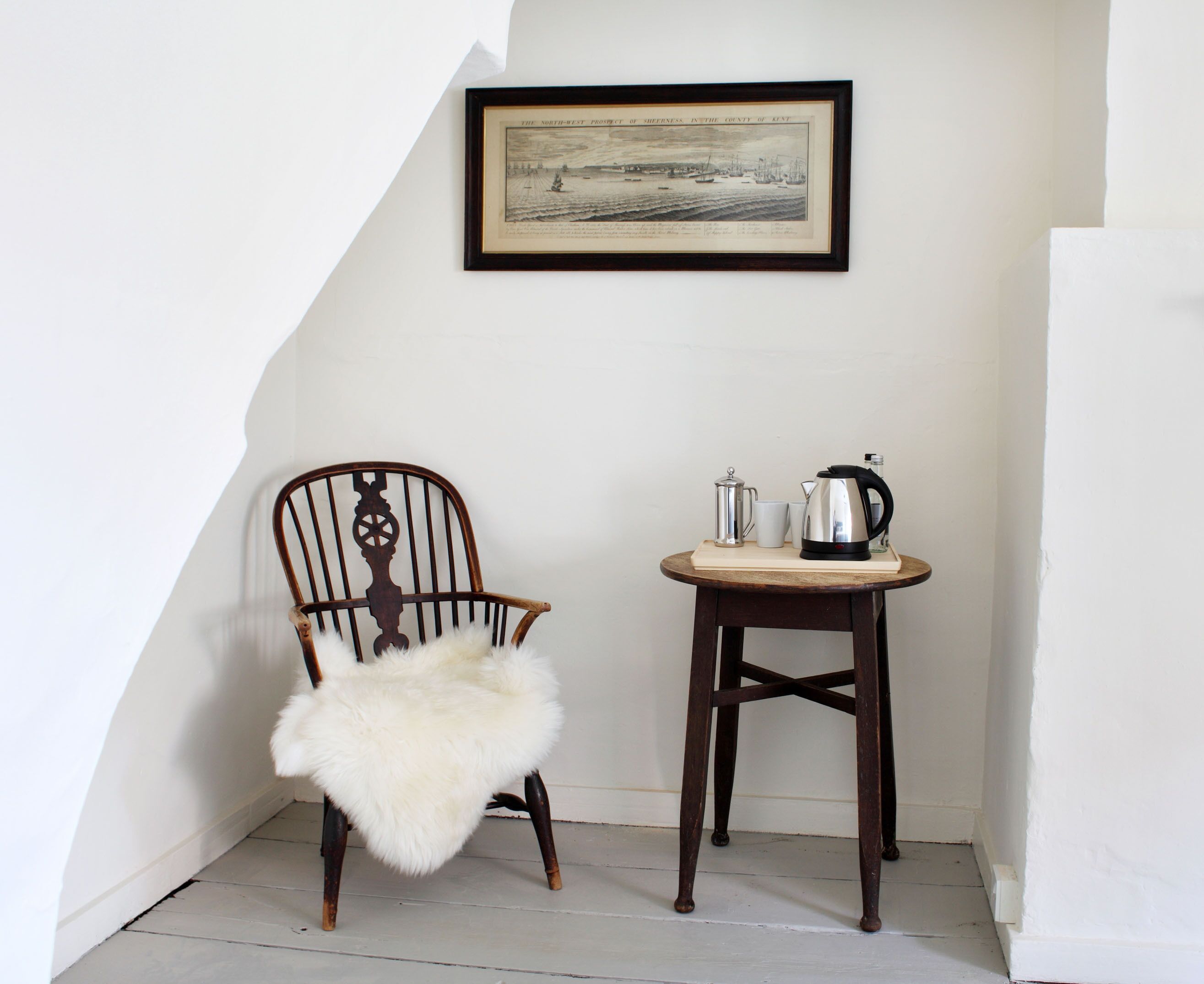 A corner room with a wooden chair, a small wooden table with a kettle, and a framed picture on the wall.