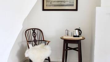 A corner room with a wooden chair, a small wooden table with a kettle, and a framed picture on the wall.