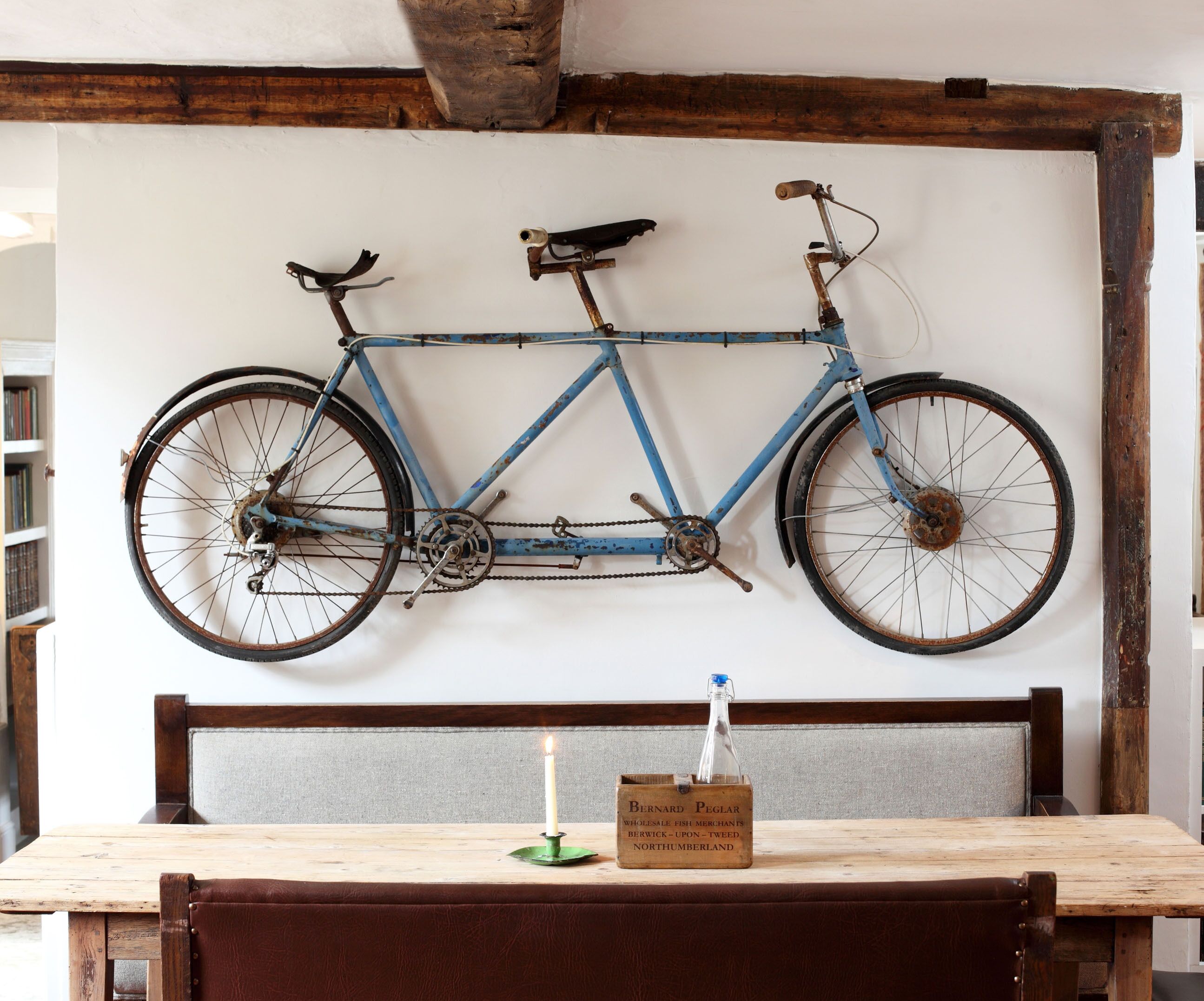 Una mesa de madera rústica con una vela y una caja de madera con la etiqueta 'BERNARD PEGLAR WHOLESALE FISH MERCHANTS' en una habitación con vigas de madera a la vista y una bicicleta colgada en la pared.