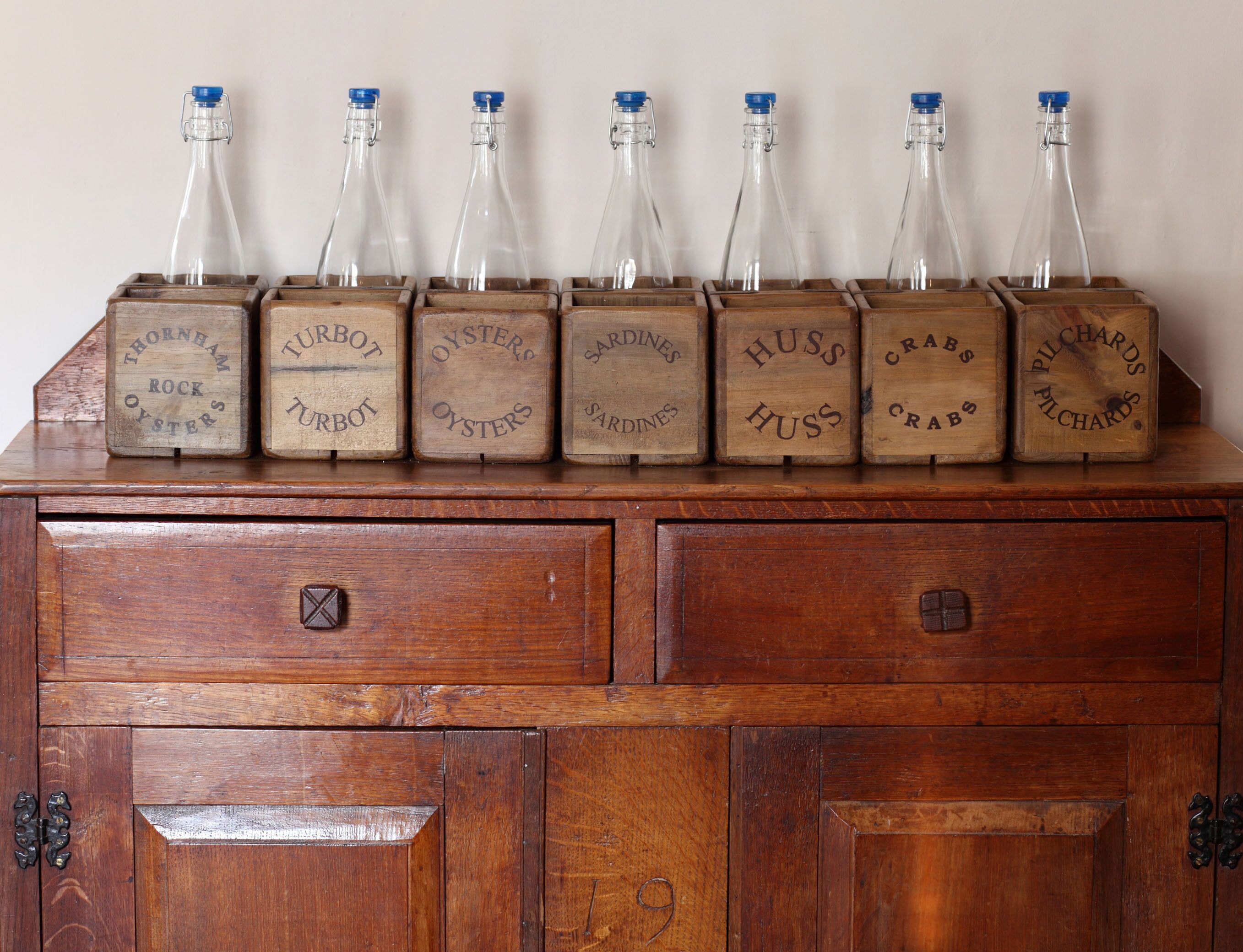 A wooden cabinet with seven glass bottles on top, each labeled with different names like "THORNHAM ROCK OYSTERS" and "HUSS."