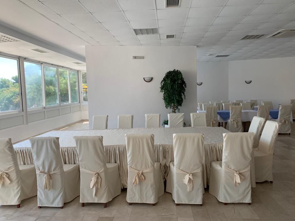 Un salón de conferencias con una larga mesa, sillas con cintas, una planta en maceta y ventanas amplias.