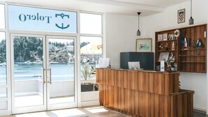 Zona de recepción con un mostrador de madera, vista al mar a través de amplios ventanales y una estantería con varios objetos, incluyendo banderas y un cuadro enmarcado.