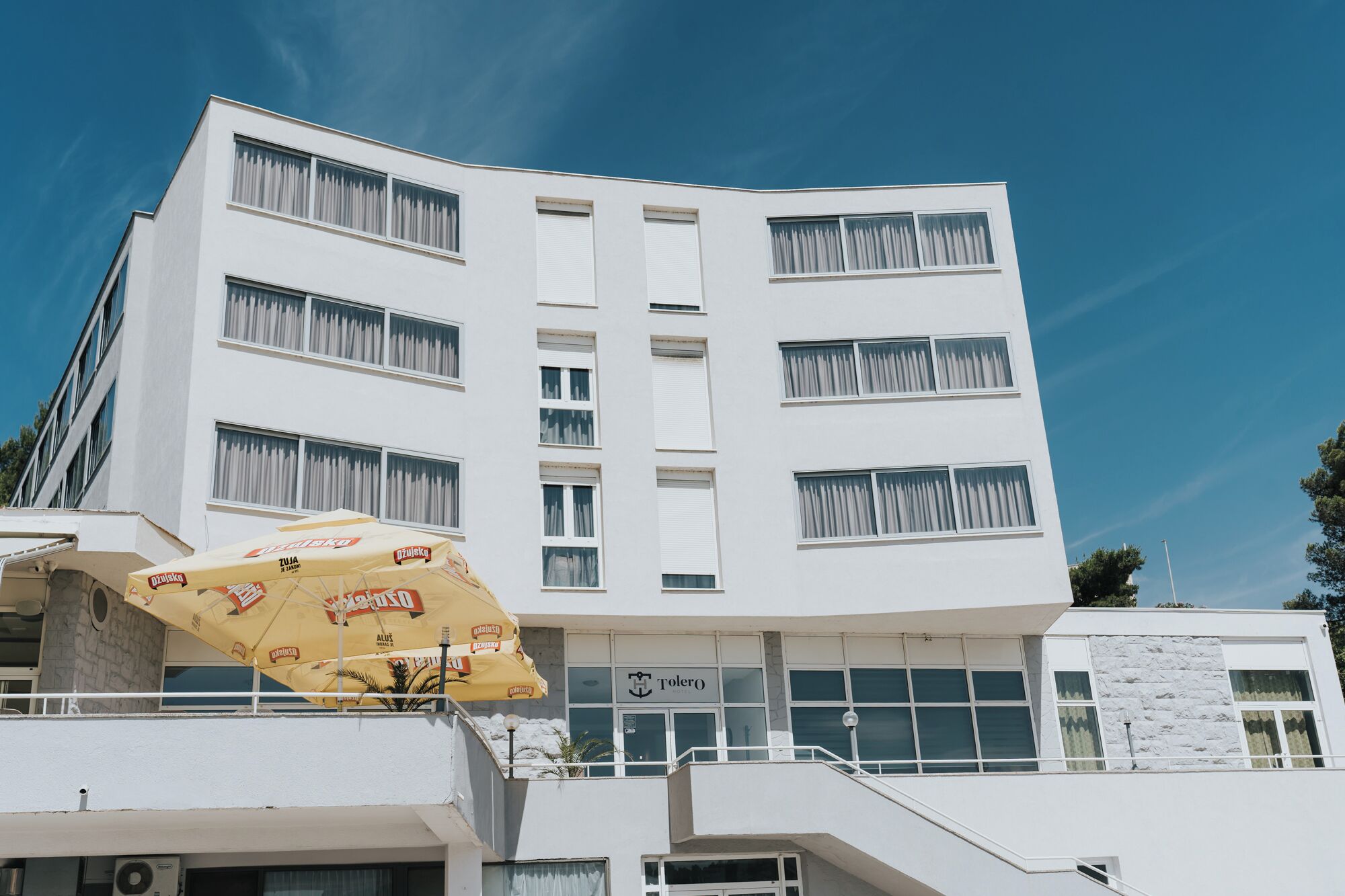 A modern hotel with a white facade, multiple windows, and a balcony with a yellow umbrella.