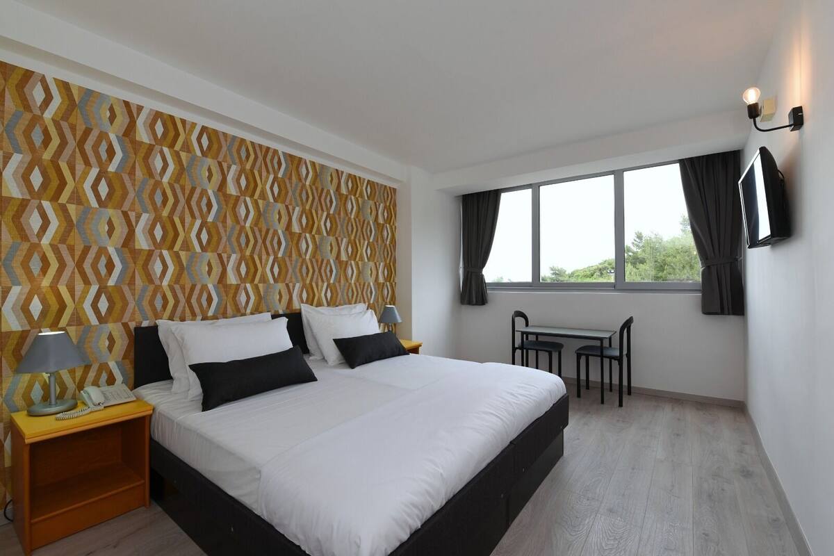 Ein Hotelzimmer mit einem großen Bett, einem Schreibtisch mit zwei Stühlen, einem an der Wand befestigten Fernseher und einer Wandtapete mit Muster am Kopfteil.