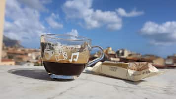 A clear glass cup of coffee with a music note design on a marble surface, a pack of almond cookies, and a cityscape in the background.